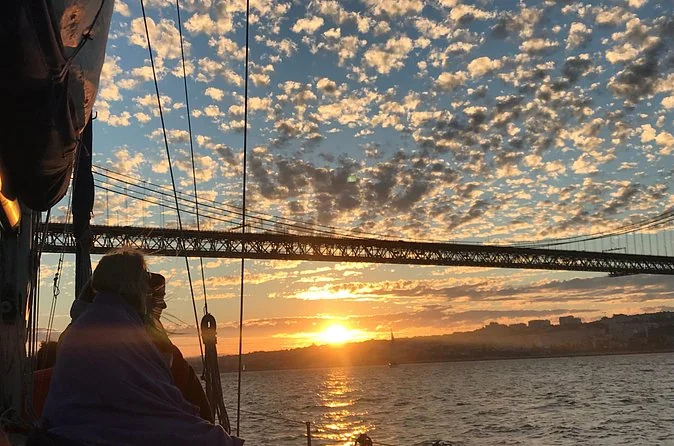 Gäste in Decken gehüllt auf einem Segelboot, während die Sonne unter der Brücke 25 de Abril untergeht